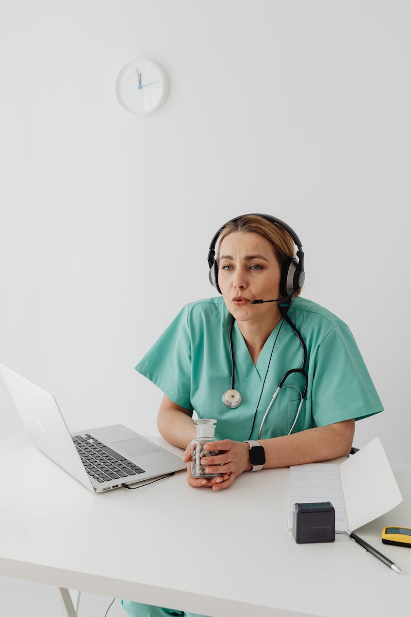 Female doctor in scrub suit conducting an online consultation using a laptop and headphones in a well-lit office.